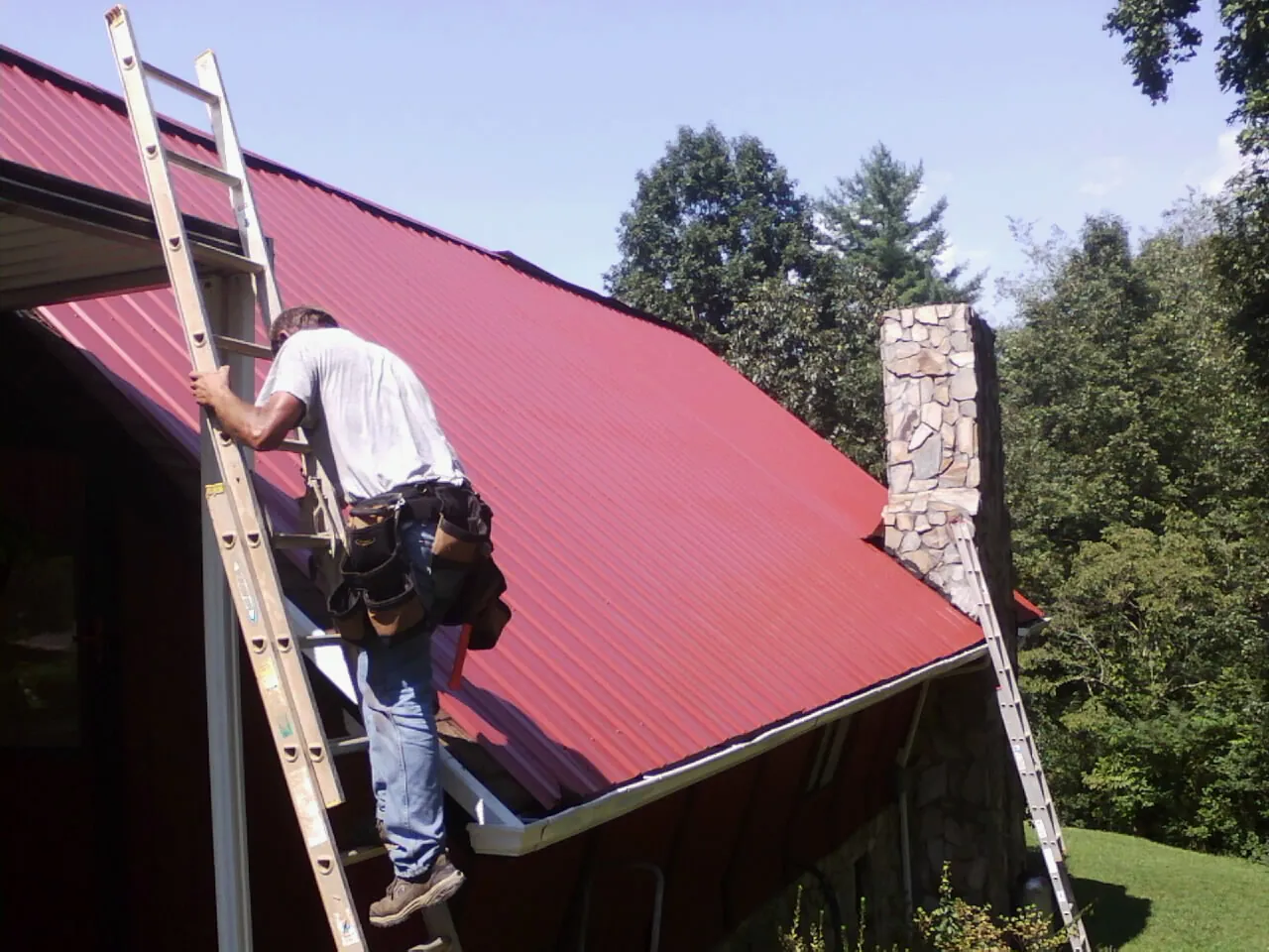 Roofer on a ladder beside a red metal roof and stone chimney — Soffit & Fascia Repair by Roofing Brothers in Black Lick